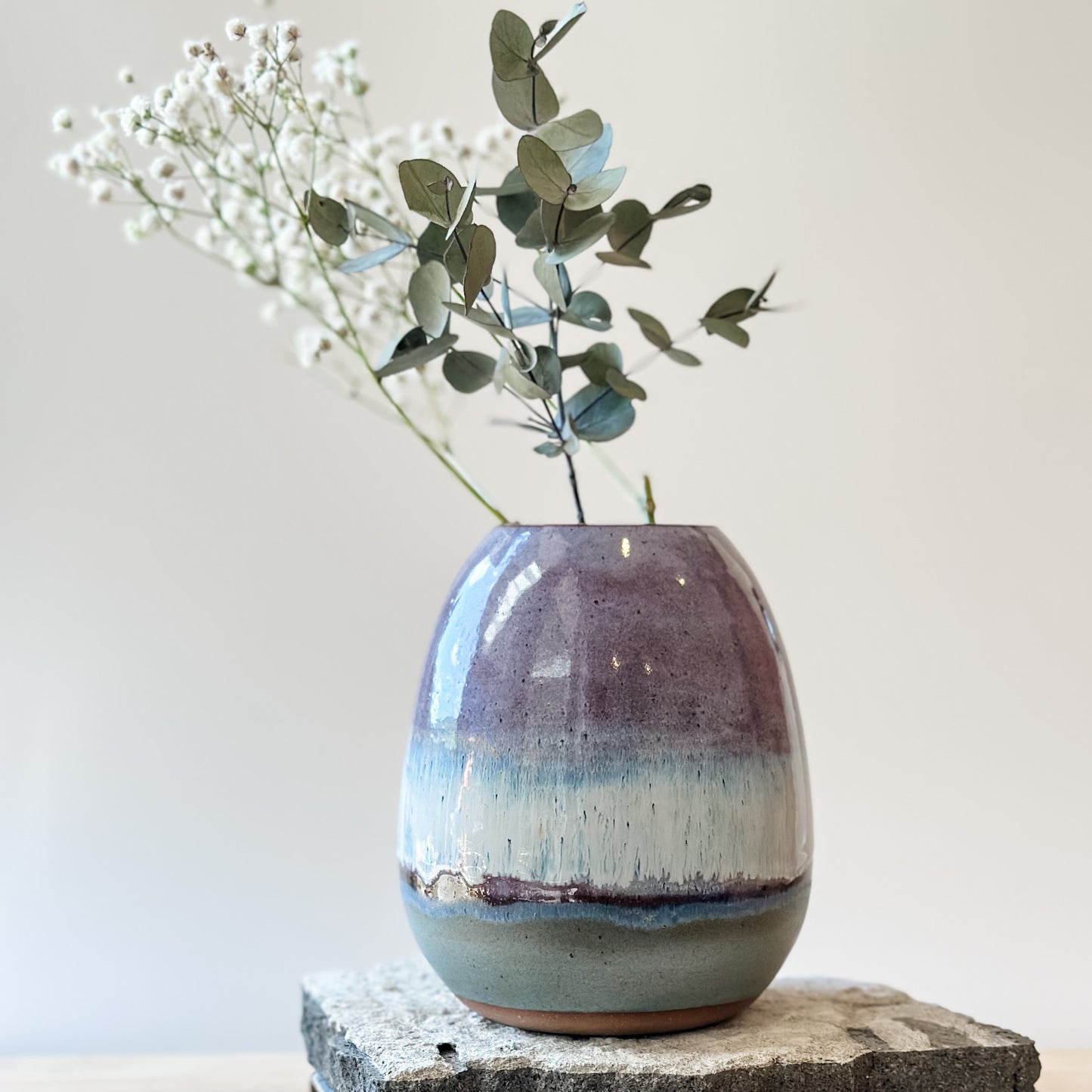 The Coastal Handmade Ceramic Round Vase sits on a tile and wooden table. The vase has a purple, white and blue glaze. The ceramic vase holds gum leaves and baby's breath flowers.
