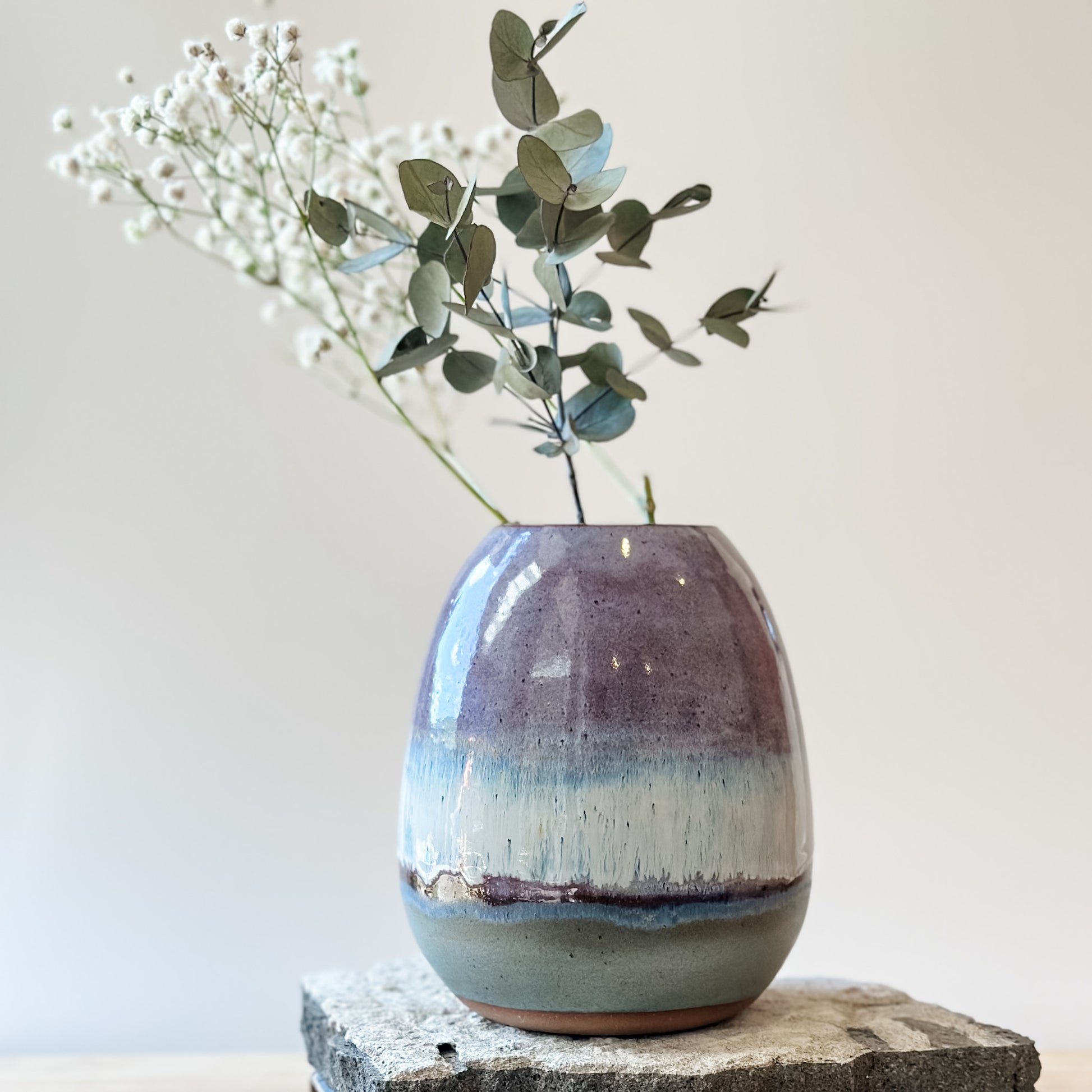 The Coastal Handmade Ceramic Round Vase sits on a tile and wooden table. The vase has a purple, white and blue glaze. The ceramic vase holds gum leaves and baby's breath flowers.