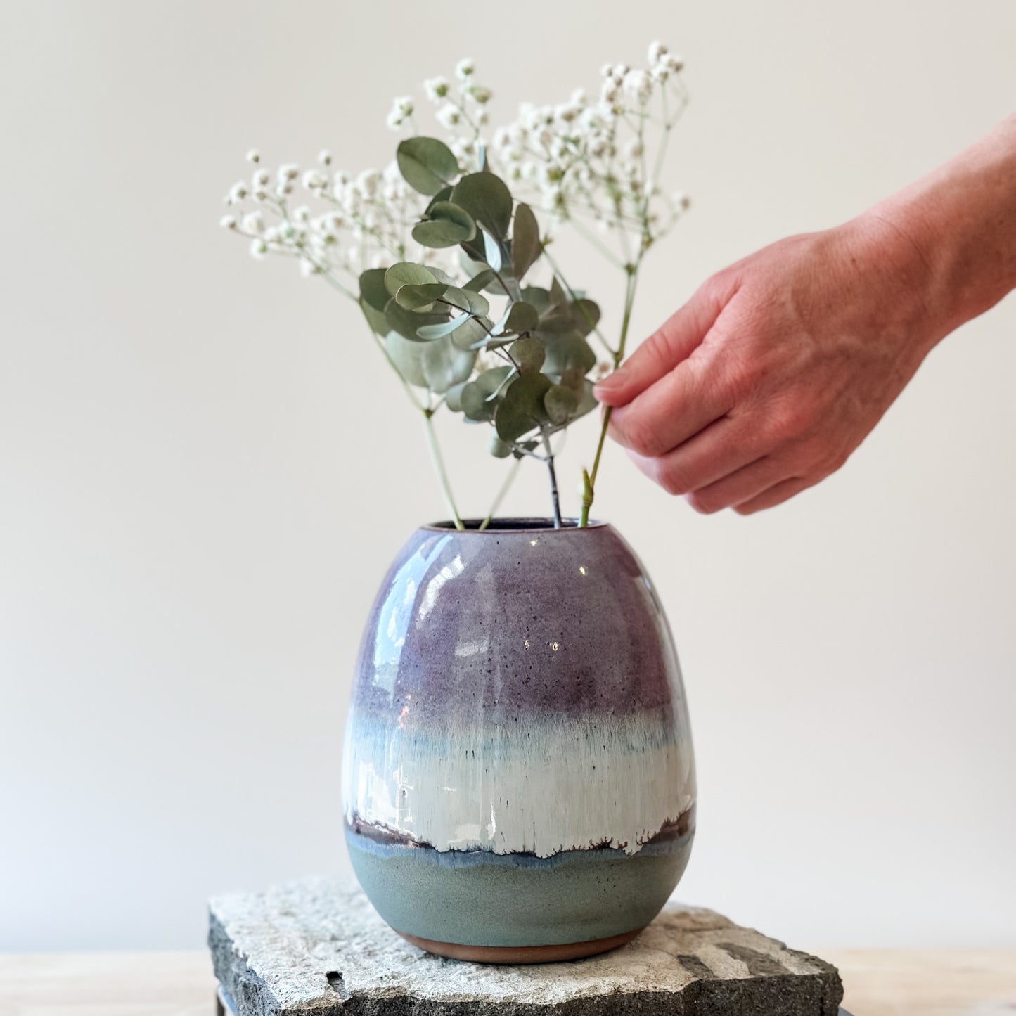 The Coastal Handmade Ceramic Round Vase sits on a tile and wooden table. The vase has a purple, white and blue glaze. A hand is arranging gum leaves and baby's breath flowers.