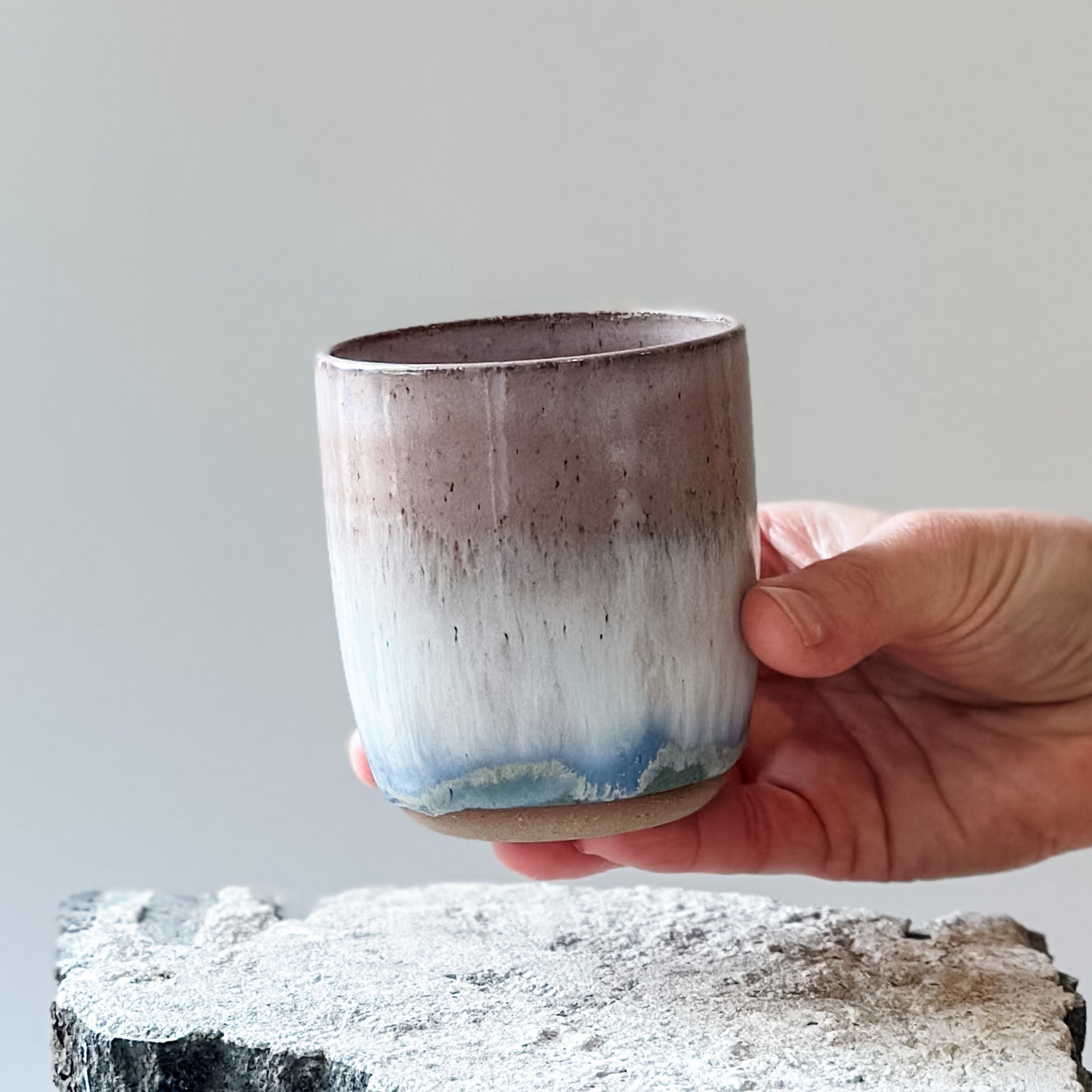A close-up view of the Coastal Handmade Ceramic Tumbler in blush pink. The handmade tumbler is being held by a hand above a tile.