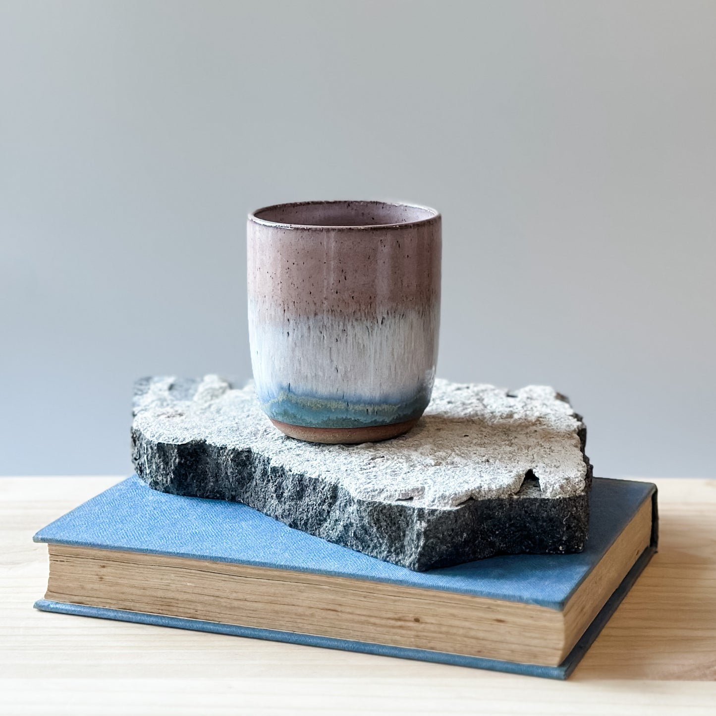 The Coastal Handmade Ceramic Tumbler in blush pink sits on a tile, a book and a wooden table.