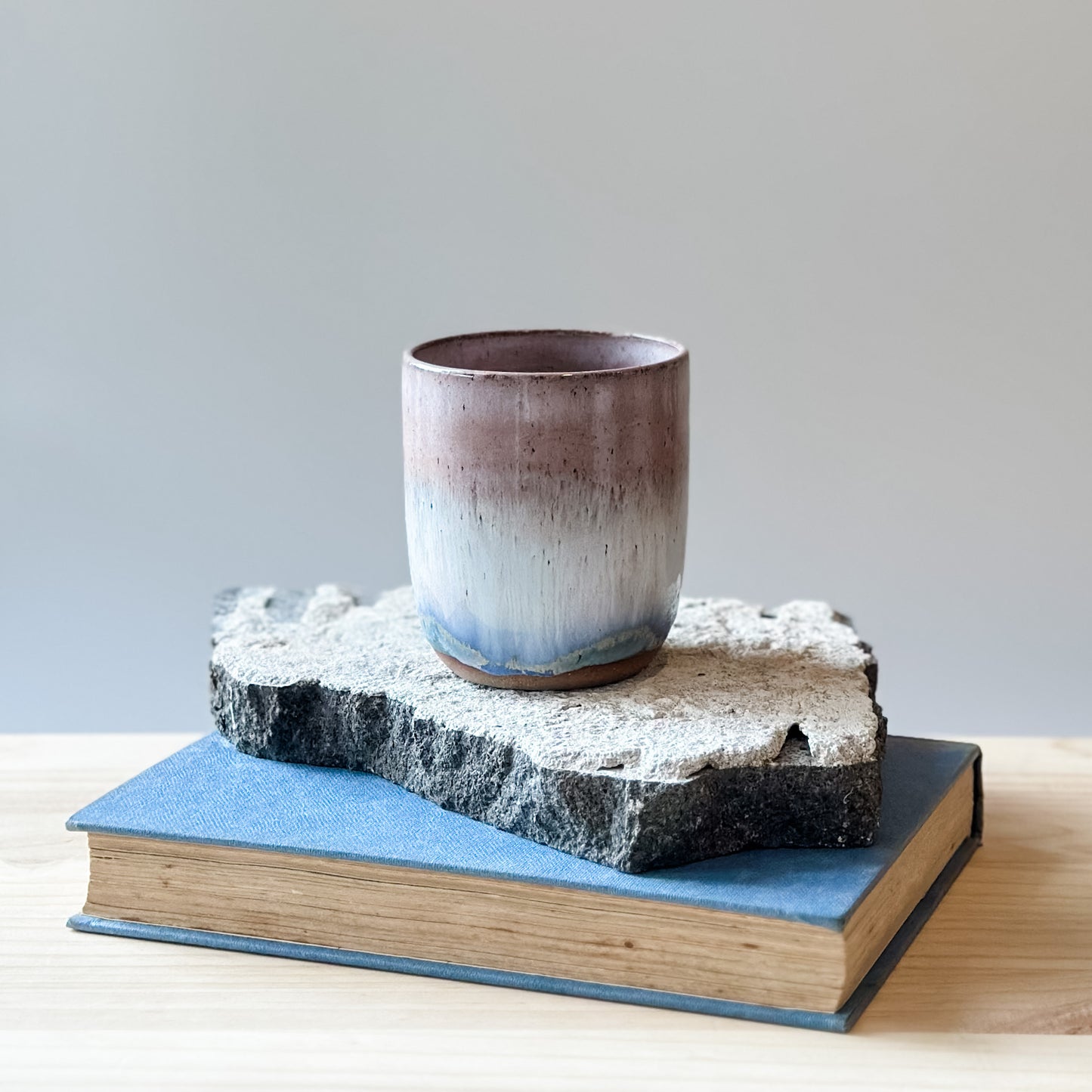 The Coastal Handmade Ceramic Tumbler in blush pink sits on a tile, a book and a wooden table.