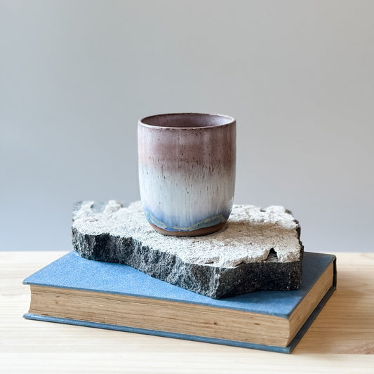 The Coastal Handmade Ceramic Tumbler in blush pink sits on a tile, a book and a wooden table.