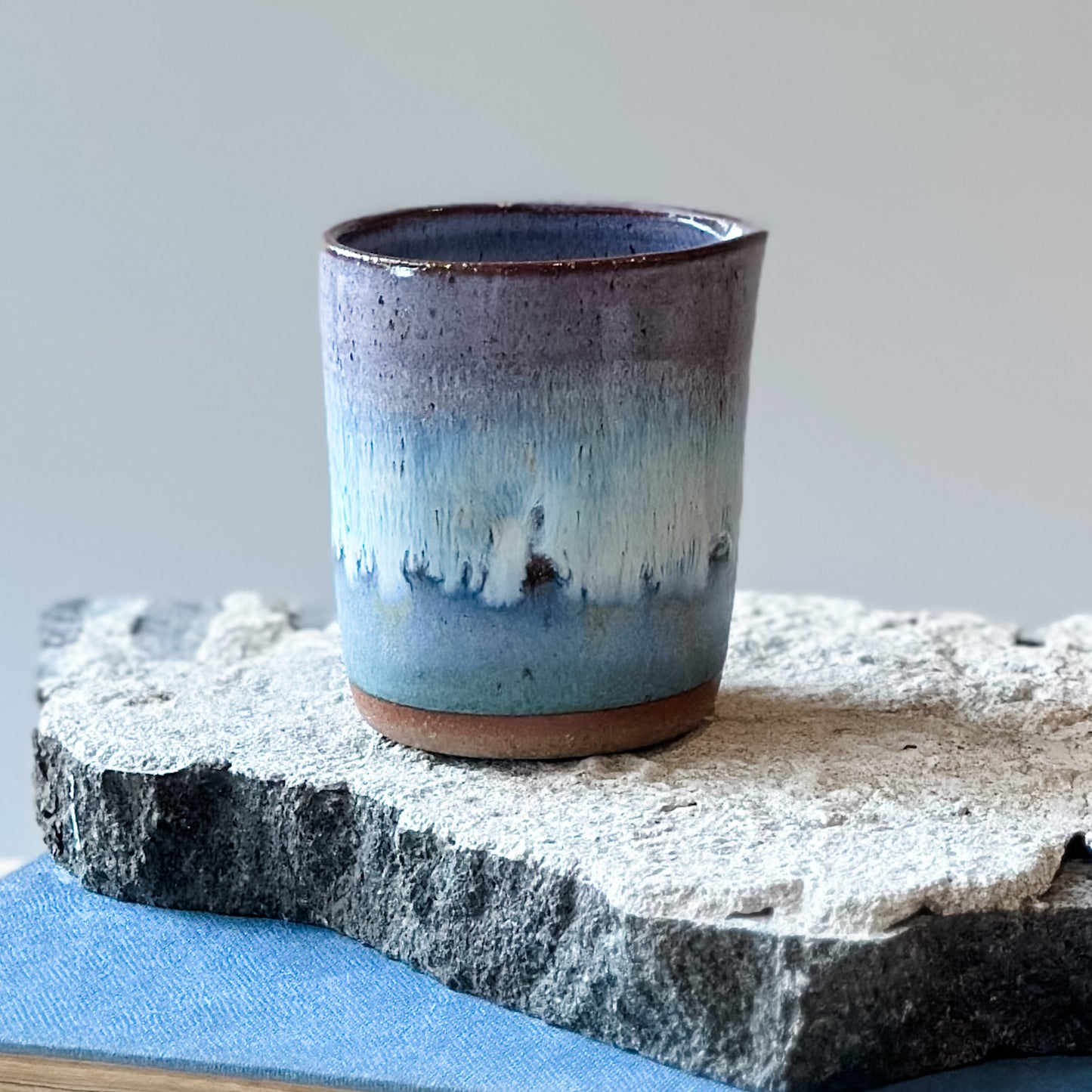 A view of the back of the Coastal Handmade Ceramic Creamer Jug. It is glazed purple, white and blue and sits on a tile and a book.