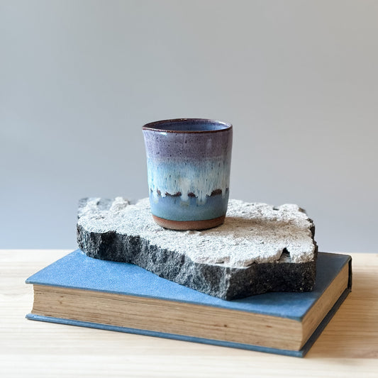 A coastal handmade ceramic creamer jug sits on a tile and a wooden table. The jug is glazed in purple, white and blue.