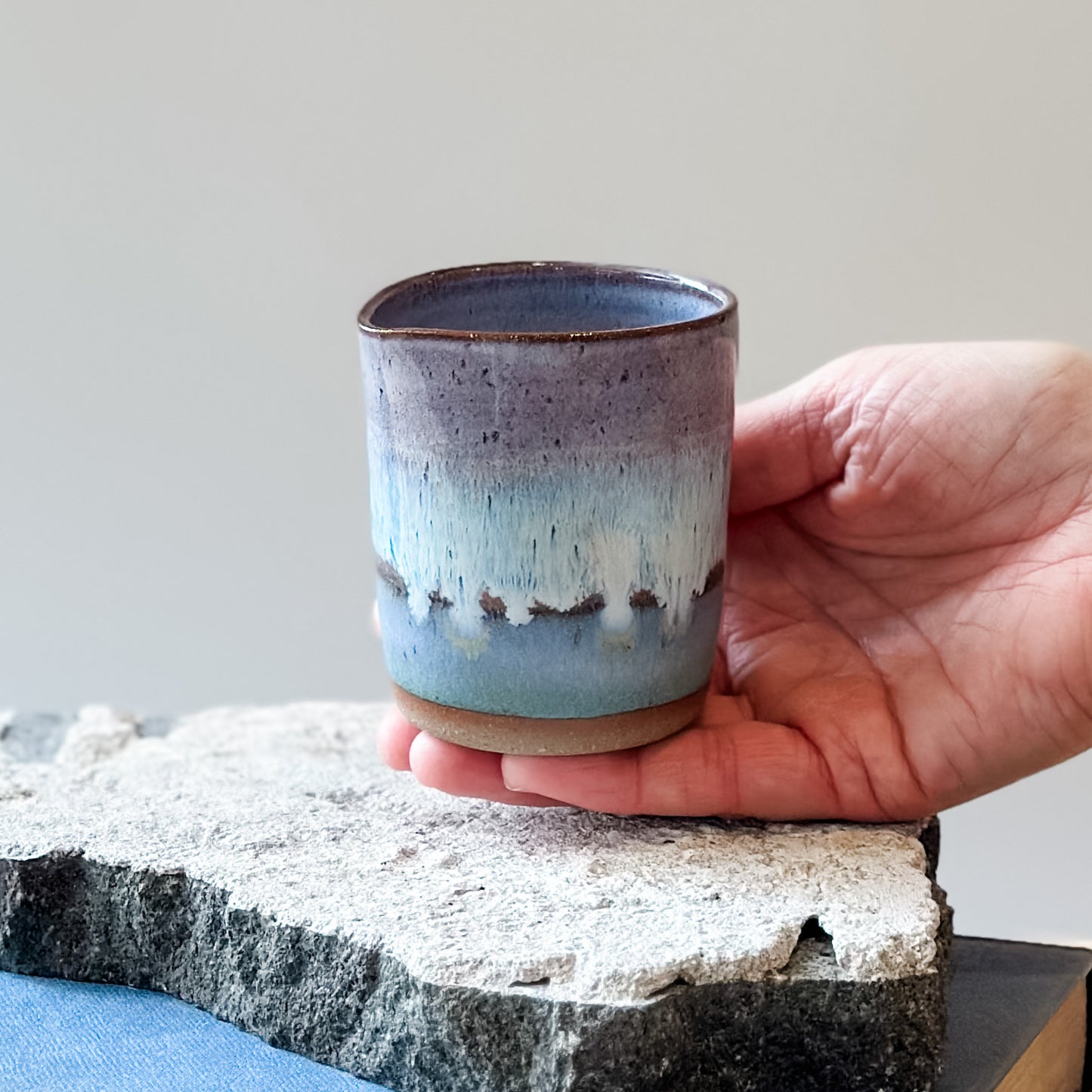 The Coastal Handmade Ceramic Creamer Jug is glazed purple, white and blue and is held above a tile.