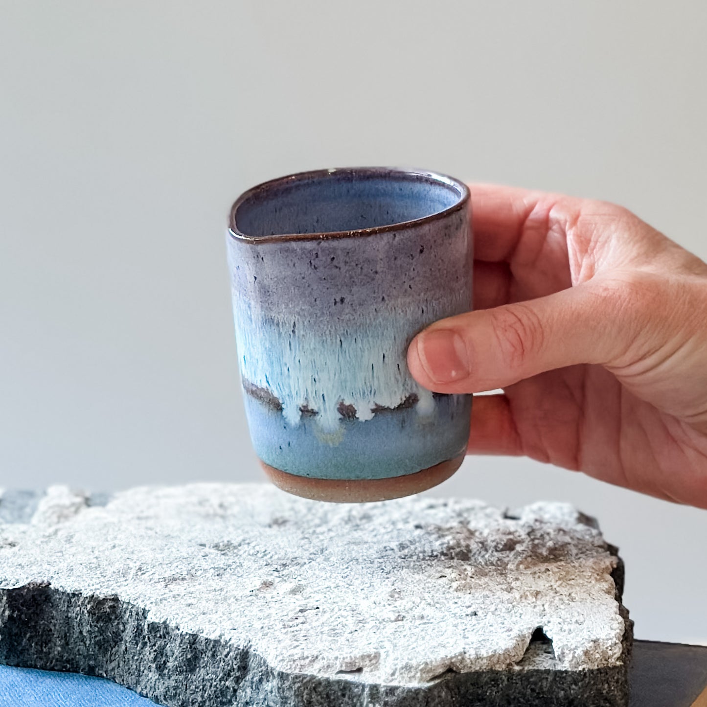 A coastal handmade ceramic creamer jug. The jug is held above a tile and glazed in purple, white and blue.