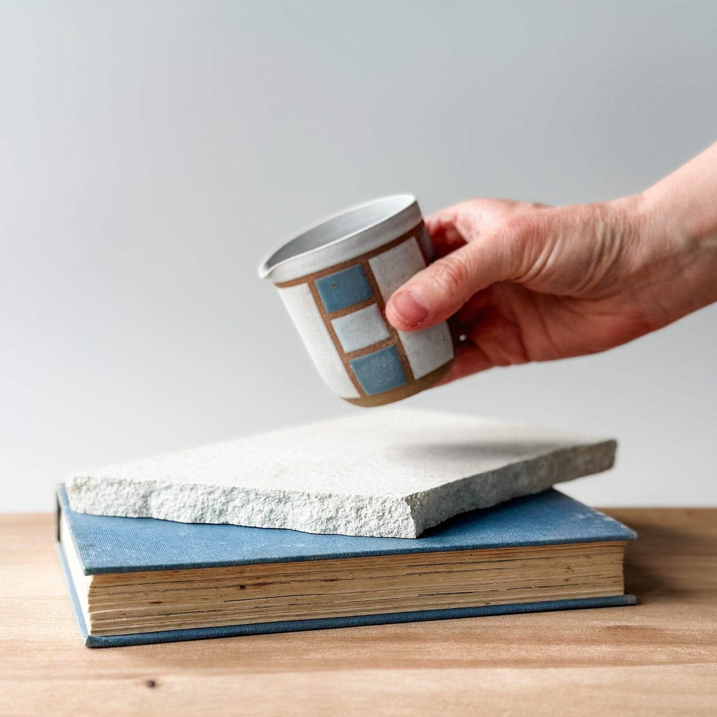 Hand holding a small ceramic cup over two stacked books on a wooden surface.