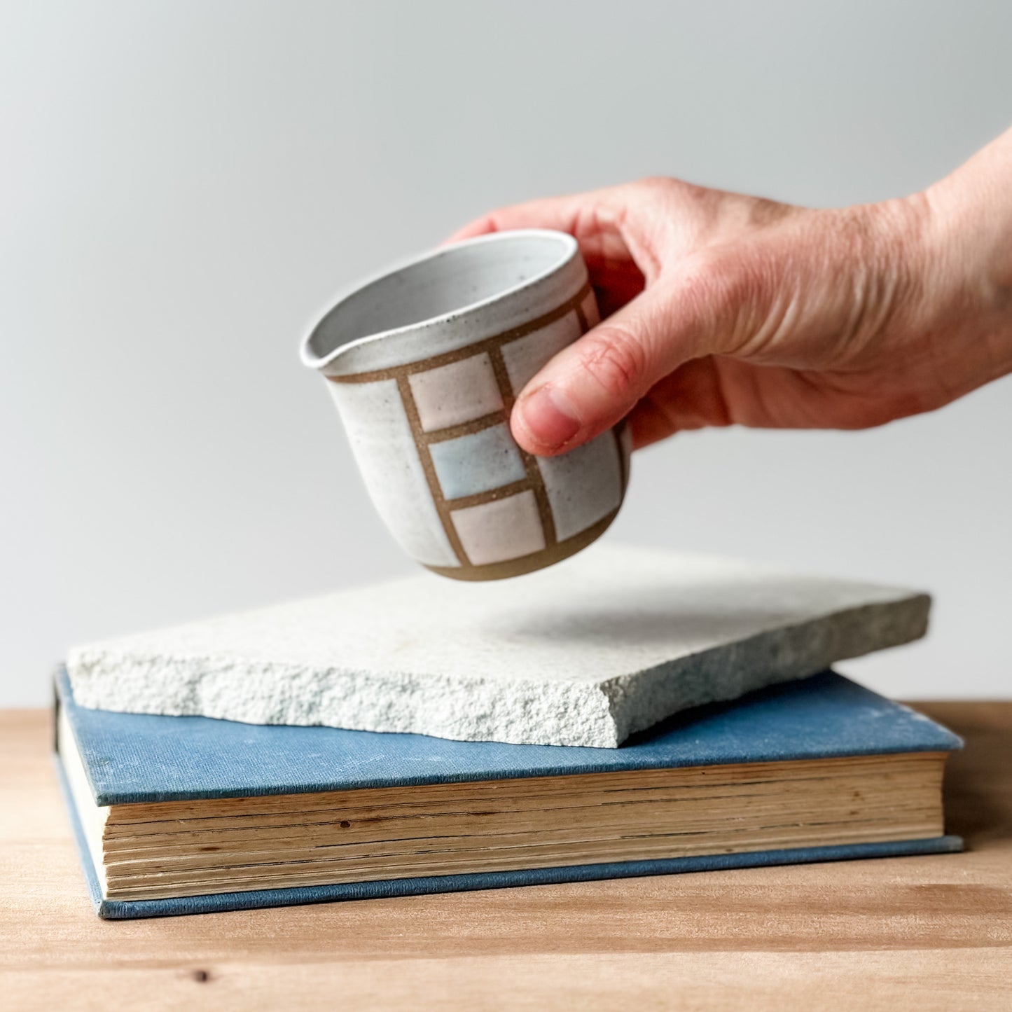 Hand holding a small ceramic cup over a stack of books on a wooden surface.
