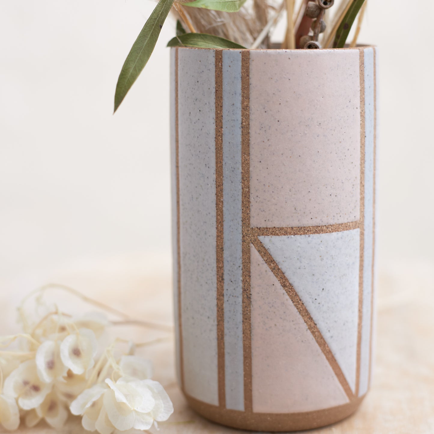 A close-up view of the Geometric Cylindrical Handmade Ceramic Vase in a pink, white and grey glaze. The handmade vase sits on a wooden stool in a coastal-styled setting. The ceramic vase displays dried, coastal flowers.