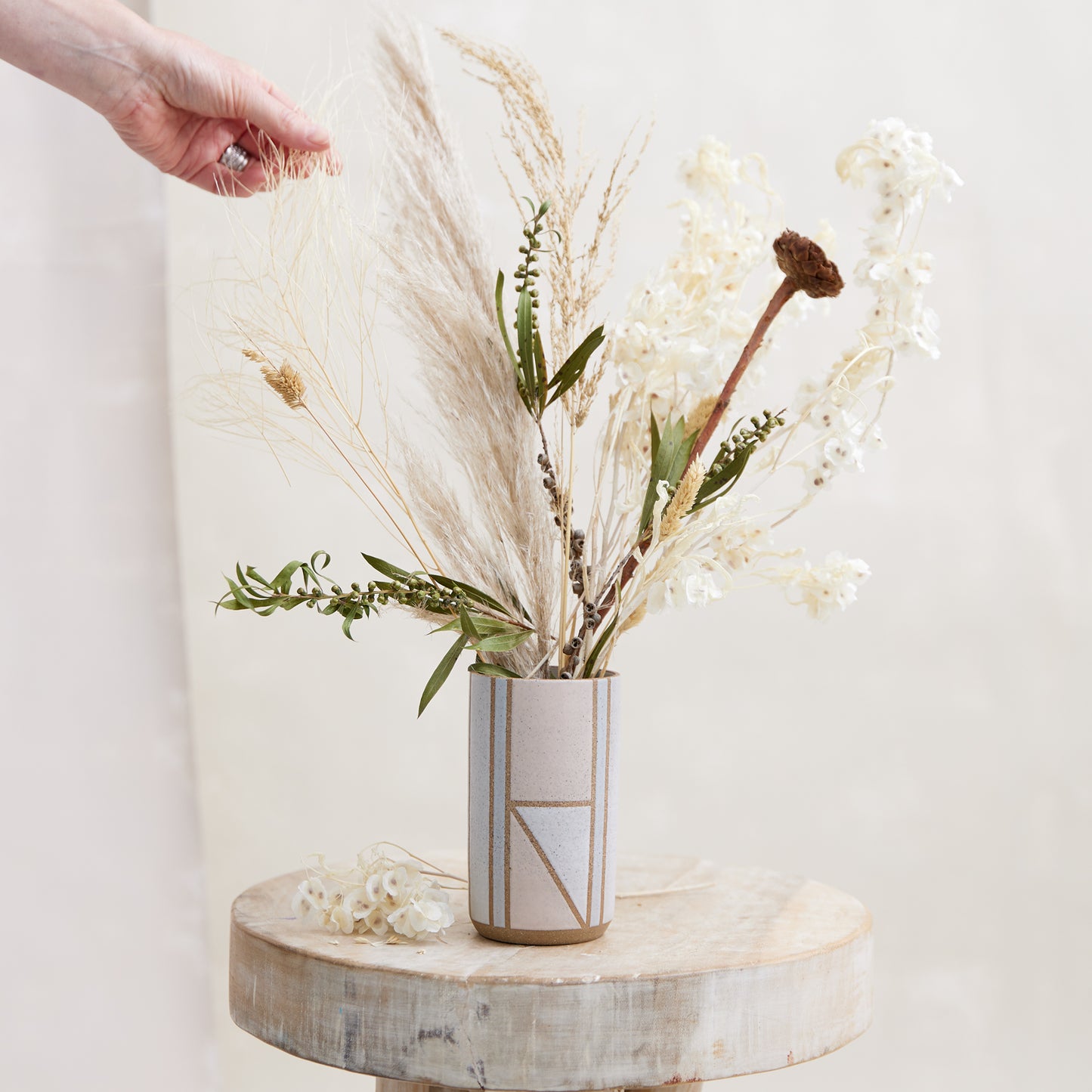 A front view of the Geometric Cylindrical Handmade Ceramic Vase in a pink, white and grey glaze. The handmade vase sits on a wooden stool in a coastal-styled setting. The ceramic vase displays dried, coastal flowers that are being adjusted by a hand.