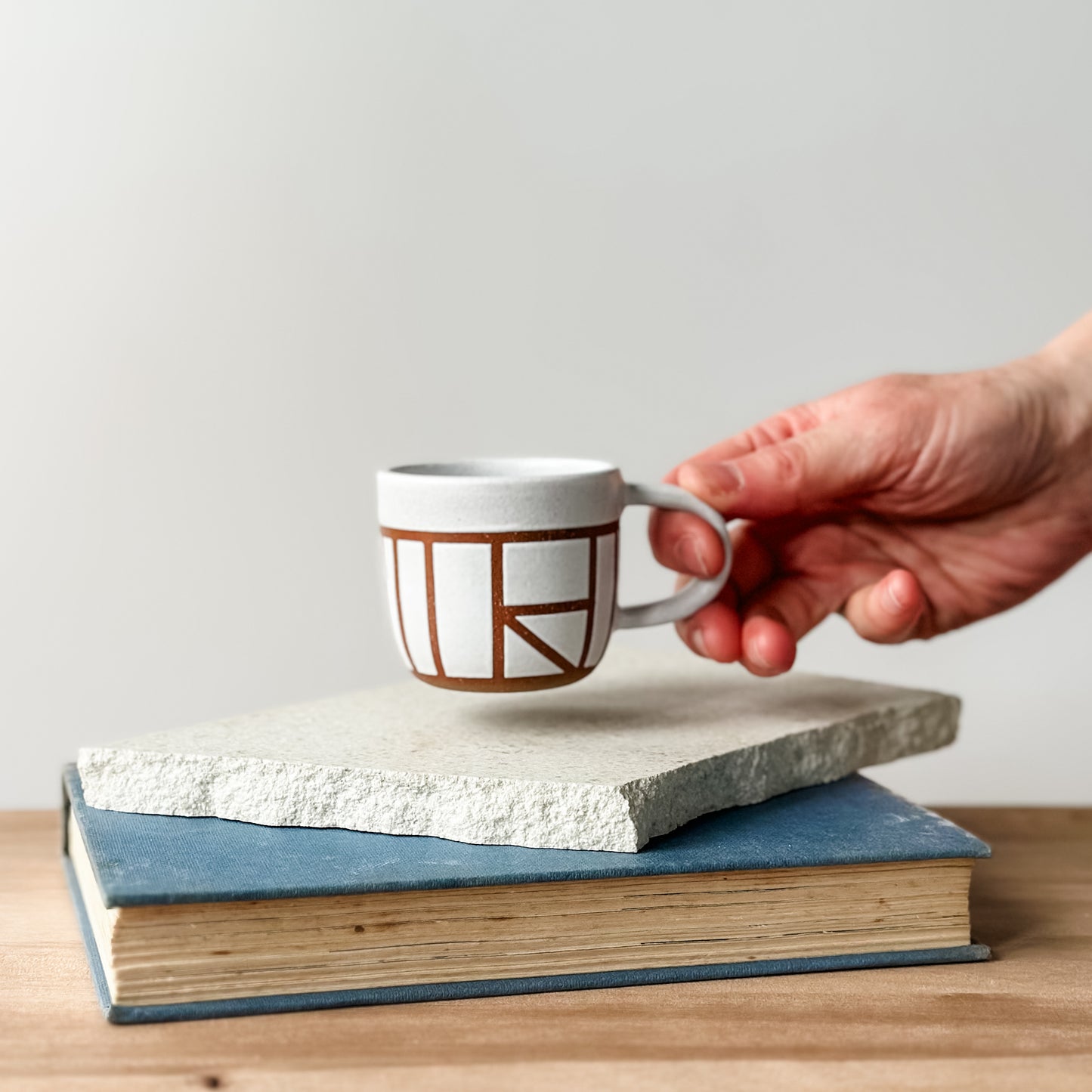 Hand holding a white mug with brown geometric design on top of two stacked books.