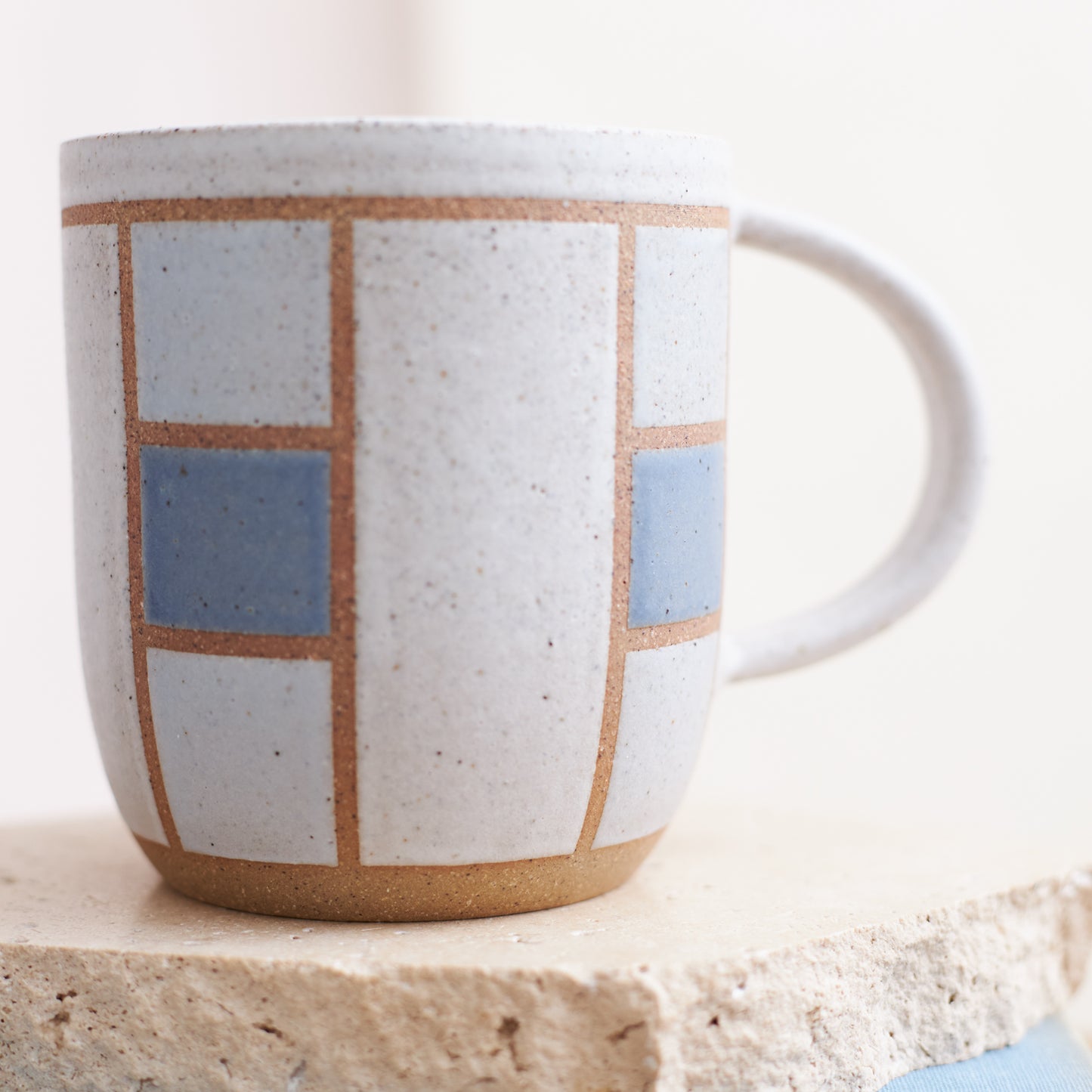 A close-up view of the Geometric Handmade Ceramic Mug in dark blue, white and grey glaze. The ceramic mug sits on a tile, book and wooden table in a coastal-styled setting.