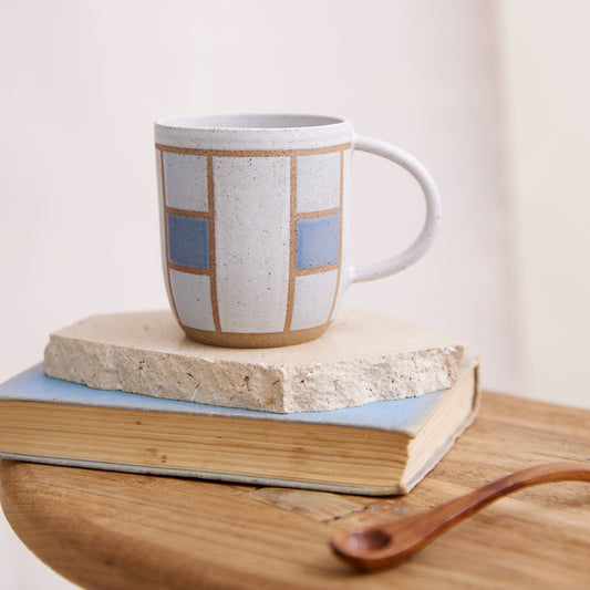 A front view of the Geometric Handmade Ceramic Mug in dark blue, white and grey glaze. The ceramic mug sits on a tile, book and wooden table in a coastal-styled setting.