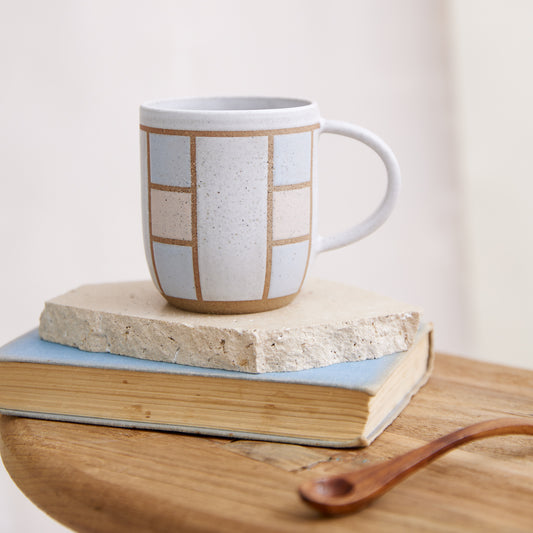 A front view of the Geometric Handmade Ceramic Mug in pink, white and grey glaze. The ceramic mug sits on a tile, book and wooden table in a coastal-styled setting.
