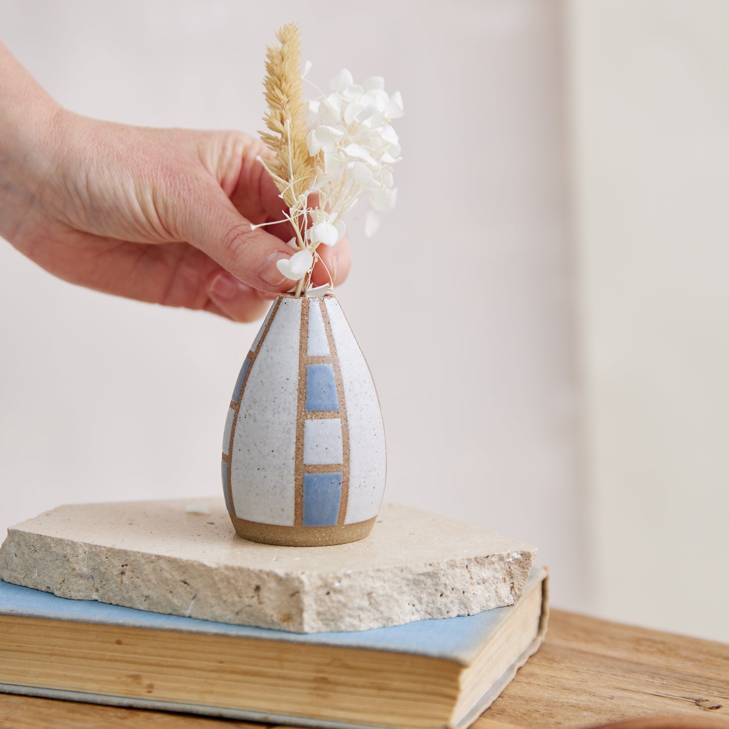 A close-up of the front of the Geometric Handmade Ceramic Mini Vase in a dark blue and white satin matte glaze. The top of the handmade vase has a tapered design. The handcrafted ceramic vase has a hand placing the coastal flowers.