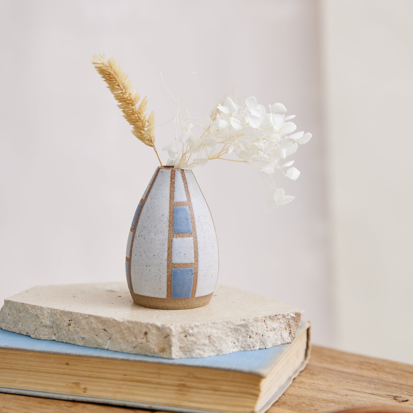 The Geometric Handmade Ceramic Mini Vase in a dark blue and white satin matte glaze. The top of the handmade vase has a tapered design. The handcrafted ceramic vase holds coastal flowers and sits on a tile, book and wooden table.