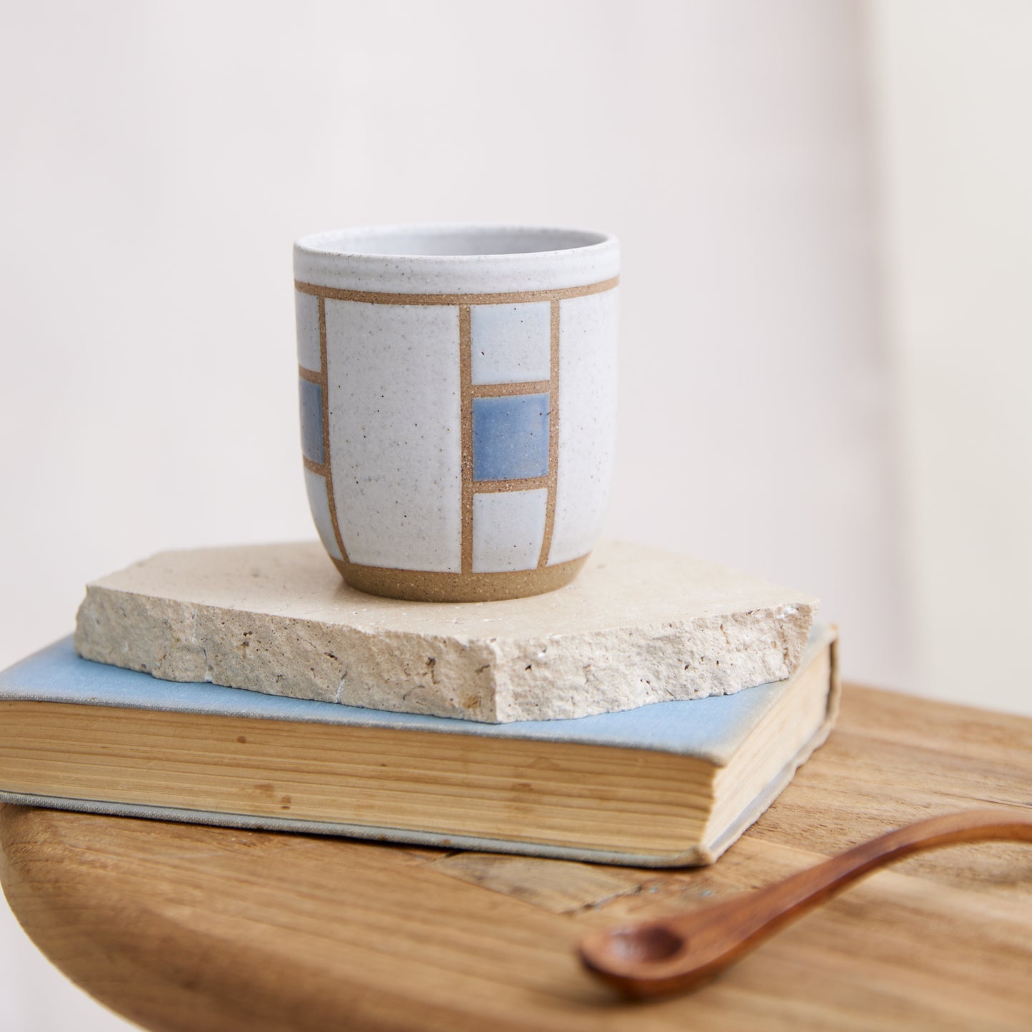 A front view of the Geometric Handmade Ceramic Tumbler in dark blue, white and grey glaze. The handmade tumbler sits on a tile, book and wooden table in a coastal-styled setting.