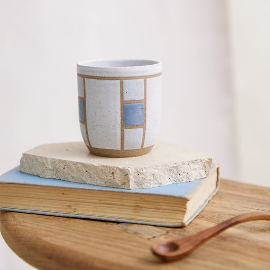 A front view of the Geometric Handmade Ceramic Tumbler in dark blue, white and grey glaze. The handmade tumbler sits on a tile, book and wooden table in a coastal-styled setting.