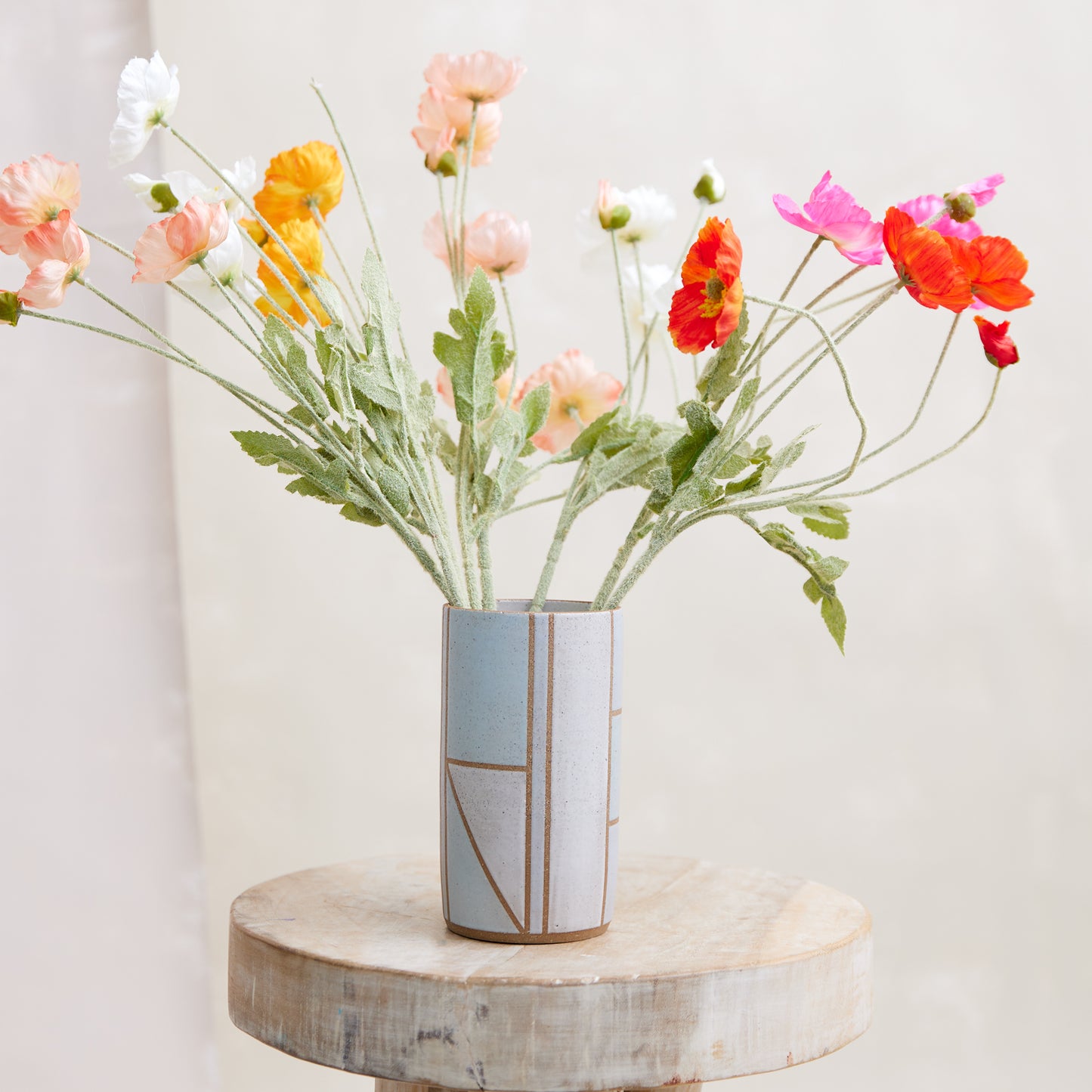 A front view of the Geometric Cylindrical Handmade Ceramic Vase in a blue, white and grey glaze. The handmade vase sits on a wooden stool in a coastal-styled setting. The ceramic vase displays fresh flowers.