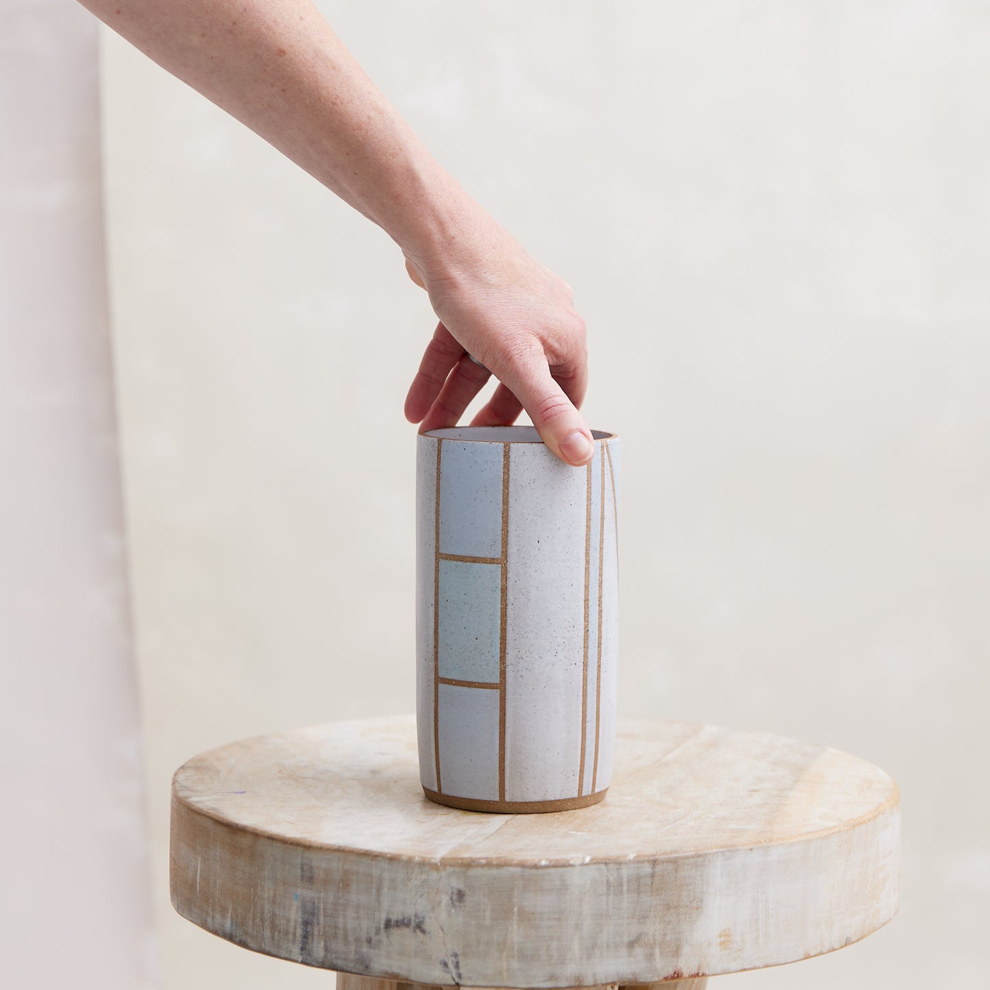 A front view of the Geometric Cylindrical Handmade Ceramic Vase in a blue, white and grey glaze. The handmade vase sits on a wooden stool in a coastal-styled setting. The ceramic vase is being adjusted by a hand.