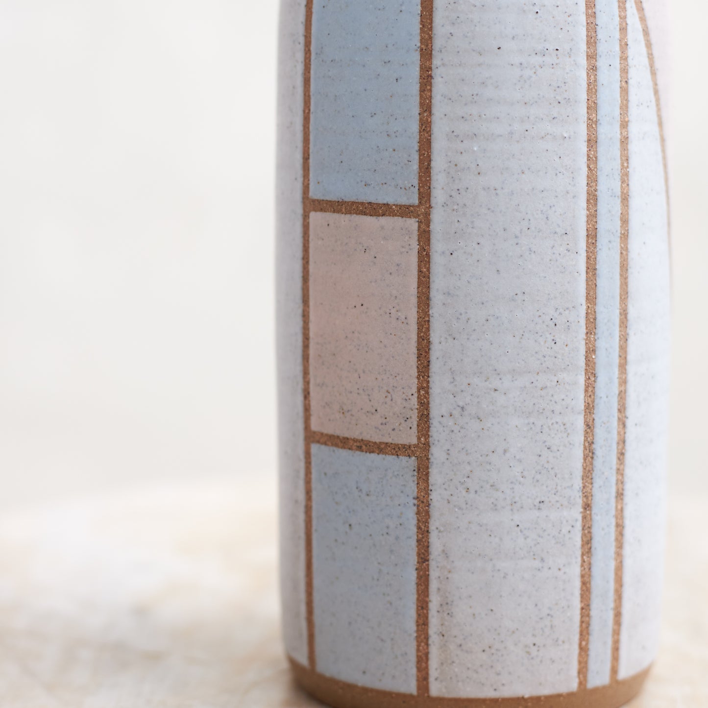 A close-up view of the Geometric Cylindrical Handmade Ceramic Vase in a pink, white and grey glaze. The handmade vase sits on a wooden stool in a coastal-styled setting.