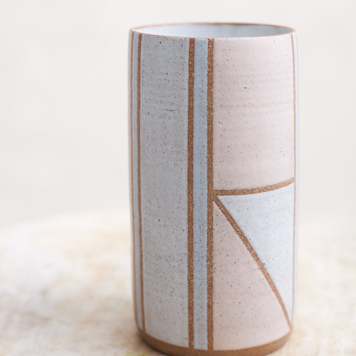 A close-up view of the Geometric Cylindrical Handmade Ceramic Vase in a pink, white and grey glaze. The handmade vase sits on a wooden stool in a coastal-styled setting.