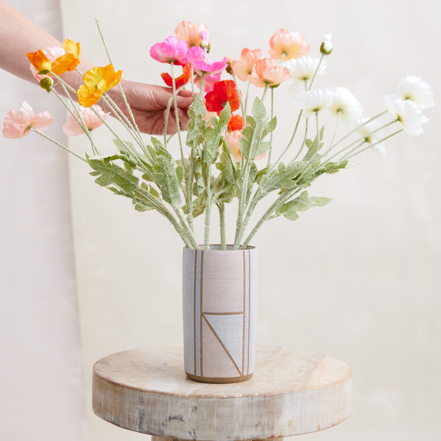A front view of the Geometric Cylindrical Handmade Ceramic Vase in a pink, white and grey glaze. The handmade vase sits on a wooden stool in a coastal-styled setting. The ceramic vase displays fresh, bright poppies that are being adjusted by a hand.