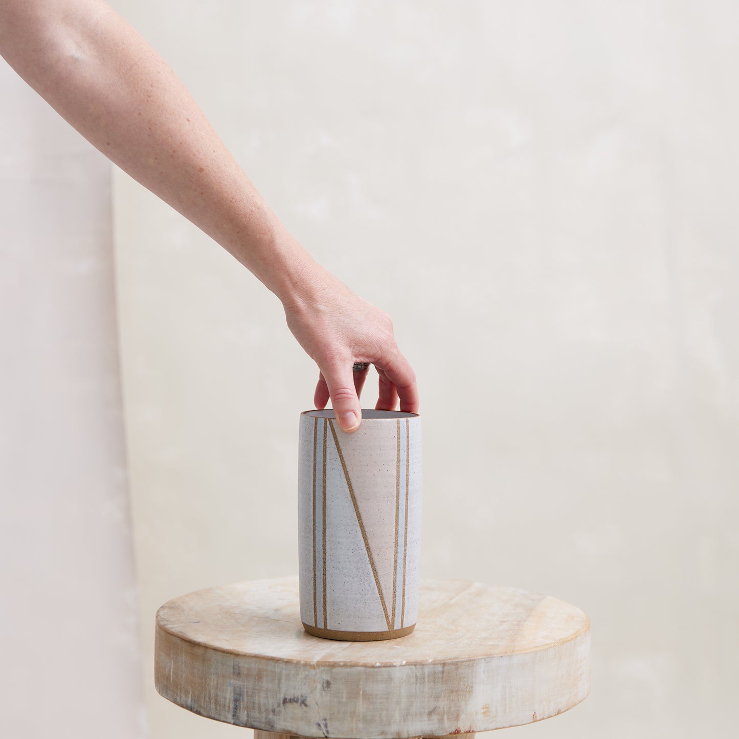 A front view of the Geometric Cylindrical Handmade Ceramic Vase in a pink, white and grey glaze. The handmade vase is placed on the wooden stool by hand. The vase is in a coastal-styled setting.