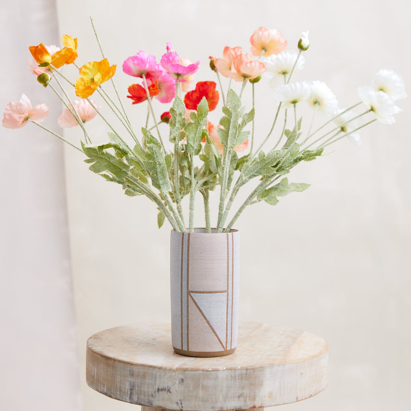 A front view of the Geometric Cylindrical Handmade Ceramic Vase in a pink, white and grey glaze. The handmade vase sits on a wooden stool in a coastal-styled setting. The ceramic vase displays fresh, bright flowers.