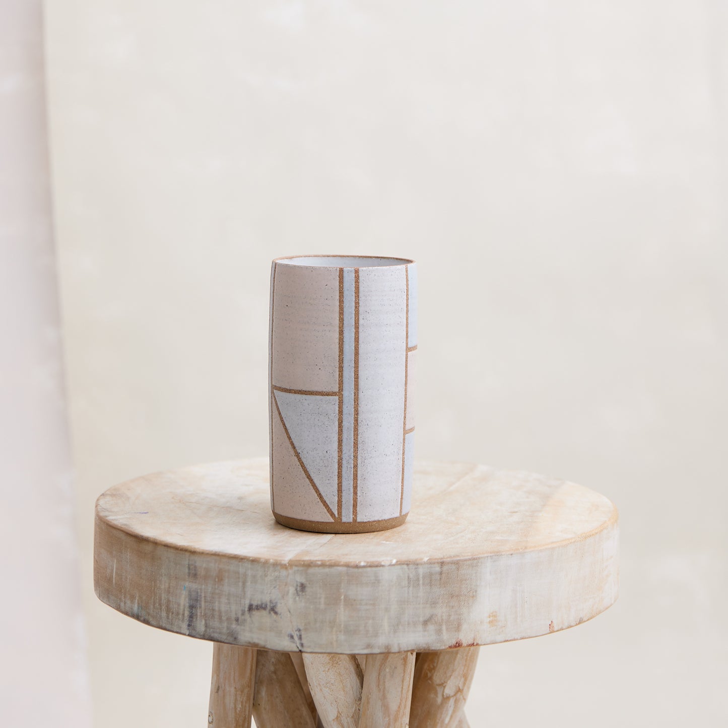 A front view of the Geometric Cylindrical Handmade Ceramic Vase in a pink, white and grey glaze. The handmade vase sits on a wooden stool in a coastal-styled setting.