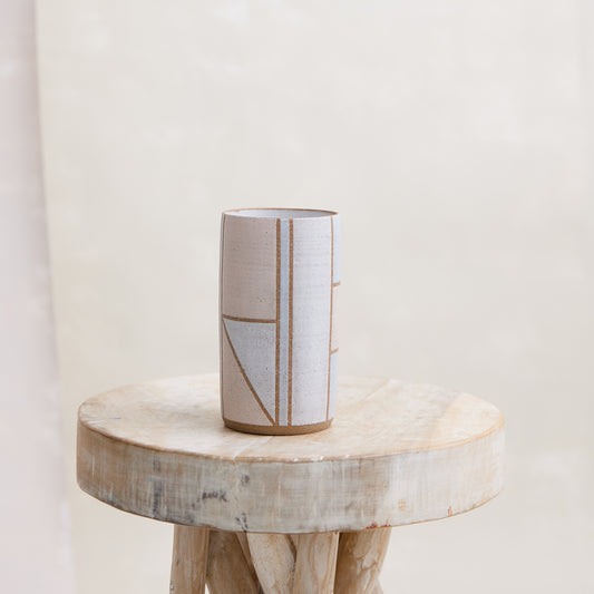 A front view of the Geometric Cylindrical Handmade Ceramic Vase in a pink, white and grey glaze. The handmade vase sits on a wooden stool in a coastal-styled setting.