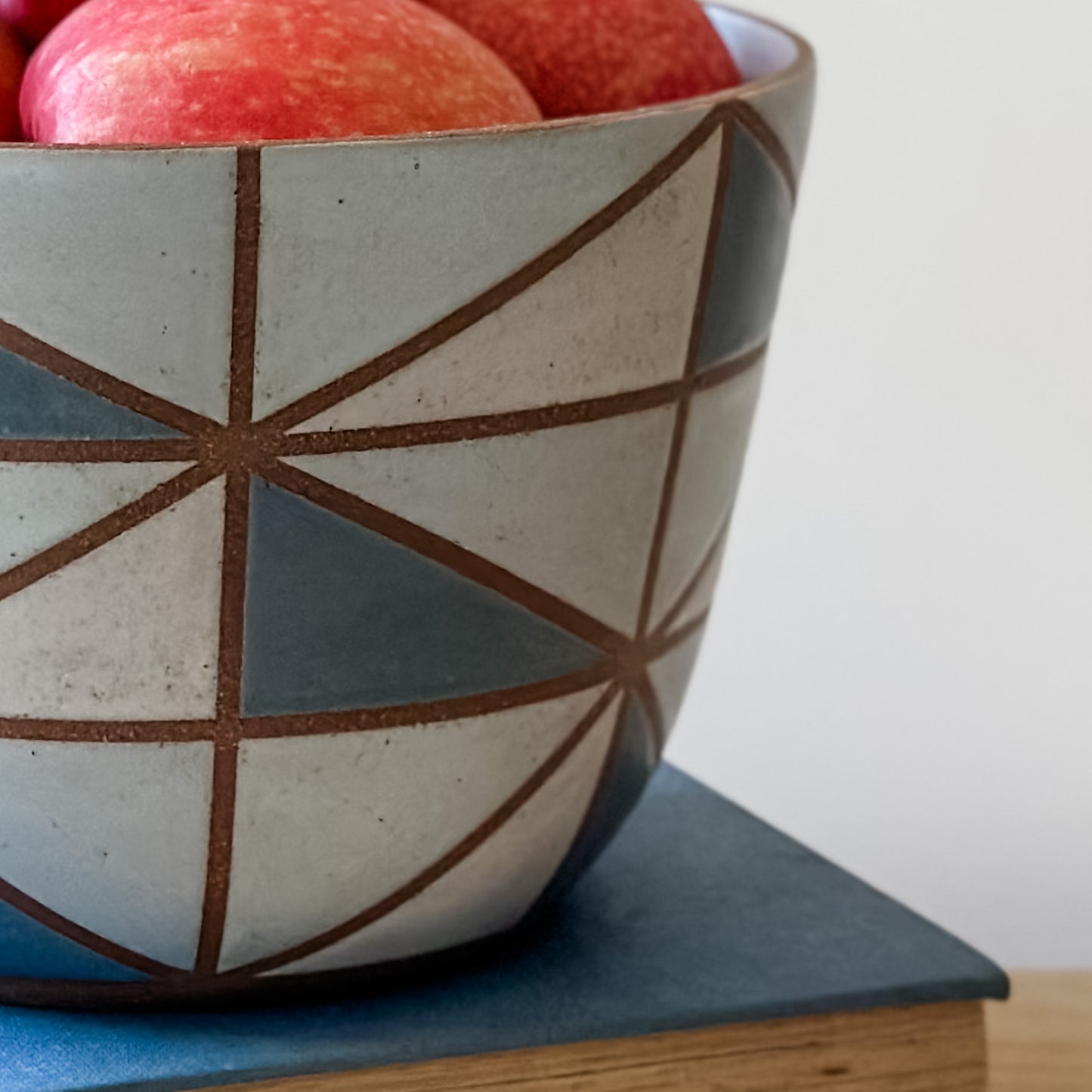 A close-up view of a geometric styled handmade ceramic bowl sitting on a book and a table. The ceramic bowl is hand-painted in dark blue, light blue, white and grey. The bowl contains fresh, red apples.