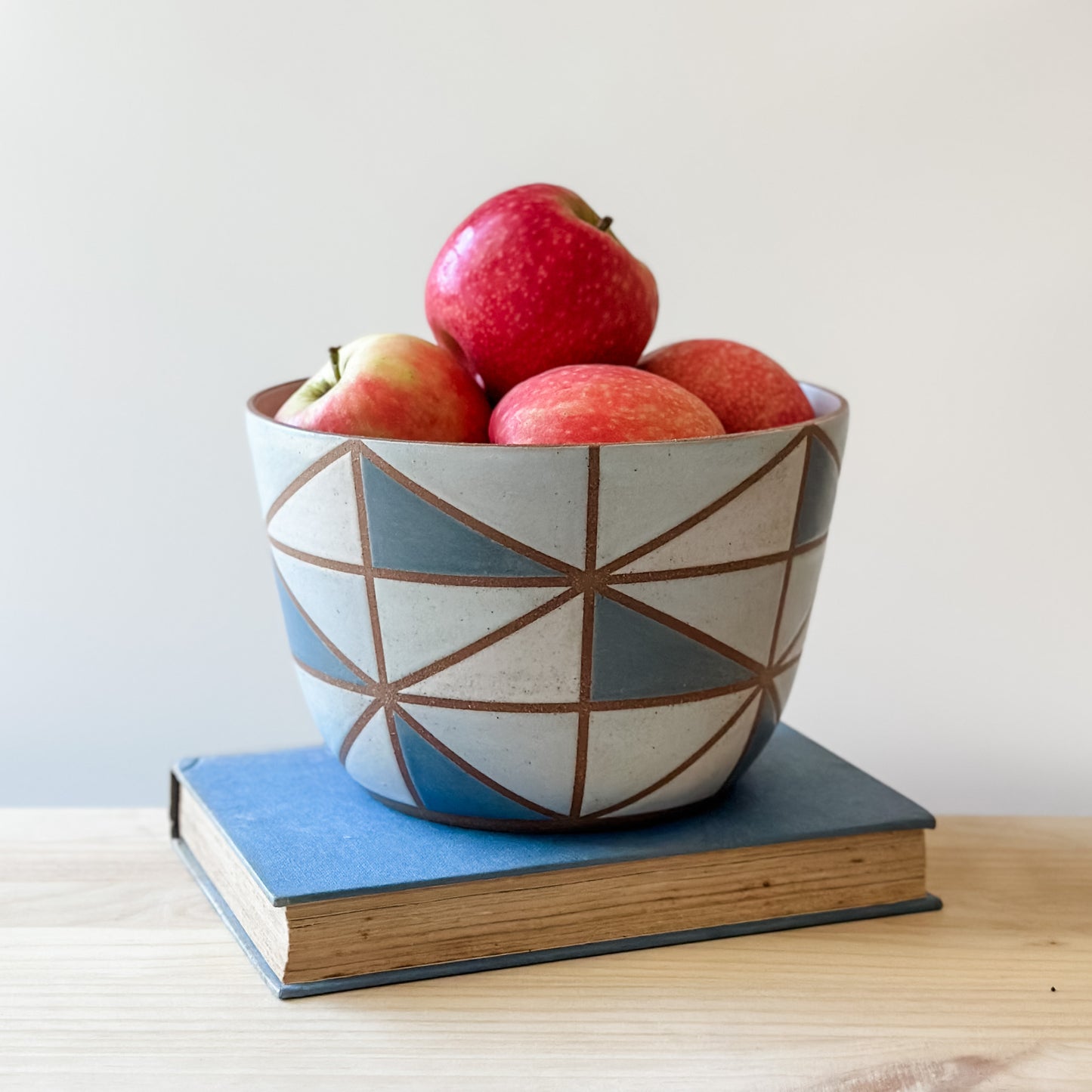 A geometric-style handmade ceramic bowl sits on a book and a table. The ceramic bowl is hand-painted in dark blue, light blue, white and grey. The bowl is holding fresh, red apples.