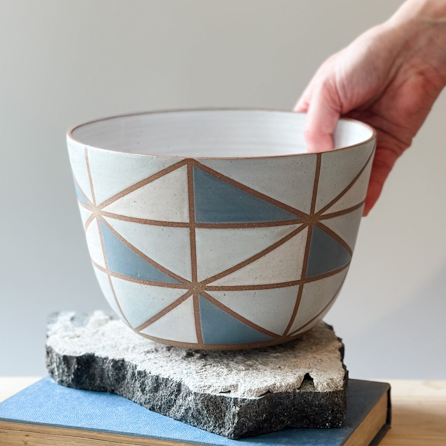 A geometric-styled handmade ceramic bowl is being placed by hand onto a tile on top of a table. The ceramic bowl is hand-painted in dark blue, light blue, white and grey.
