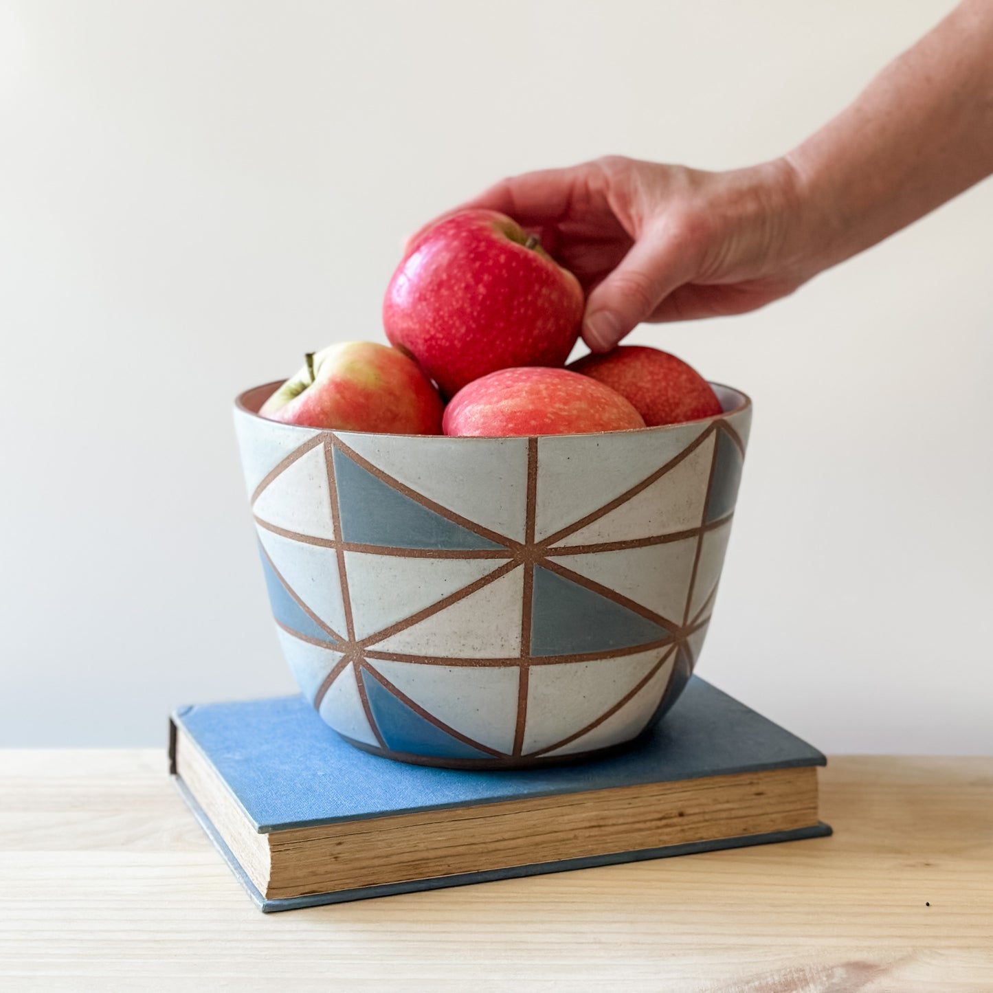 A geometric-styled handmade ceramic bowl sits on a book on top of a table. The ceramic bowl is hand-painted in dark blue, light blue, white and grey. A hand is placing fresh, red apples into the bowl.