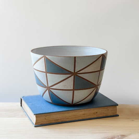 A geometric-style handmade ceramic bowl sits on a book and a table. The ceramic bowl is hand-painted in dark blue, light blue, white and grey.