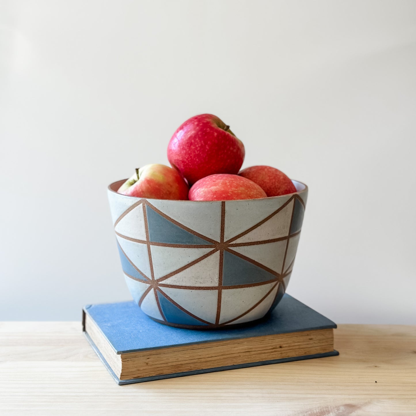 A geometric-styled handmade ceramic bowl sits on a book on top of a table. The ceramic bowl is hand-painted in dark blue, light blue, white and grey. The ceramic bowl is holding fresh, red apples.