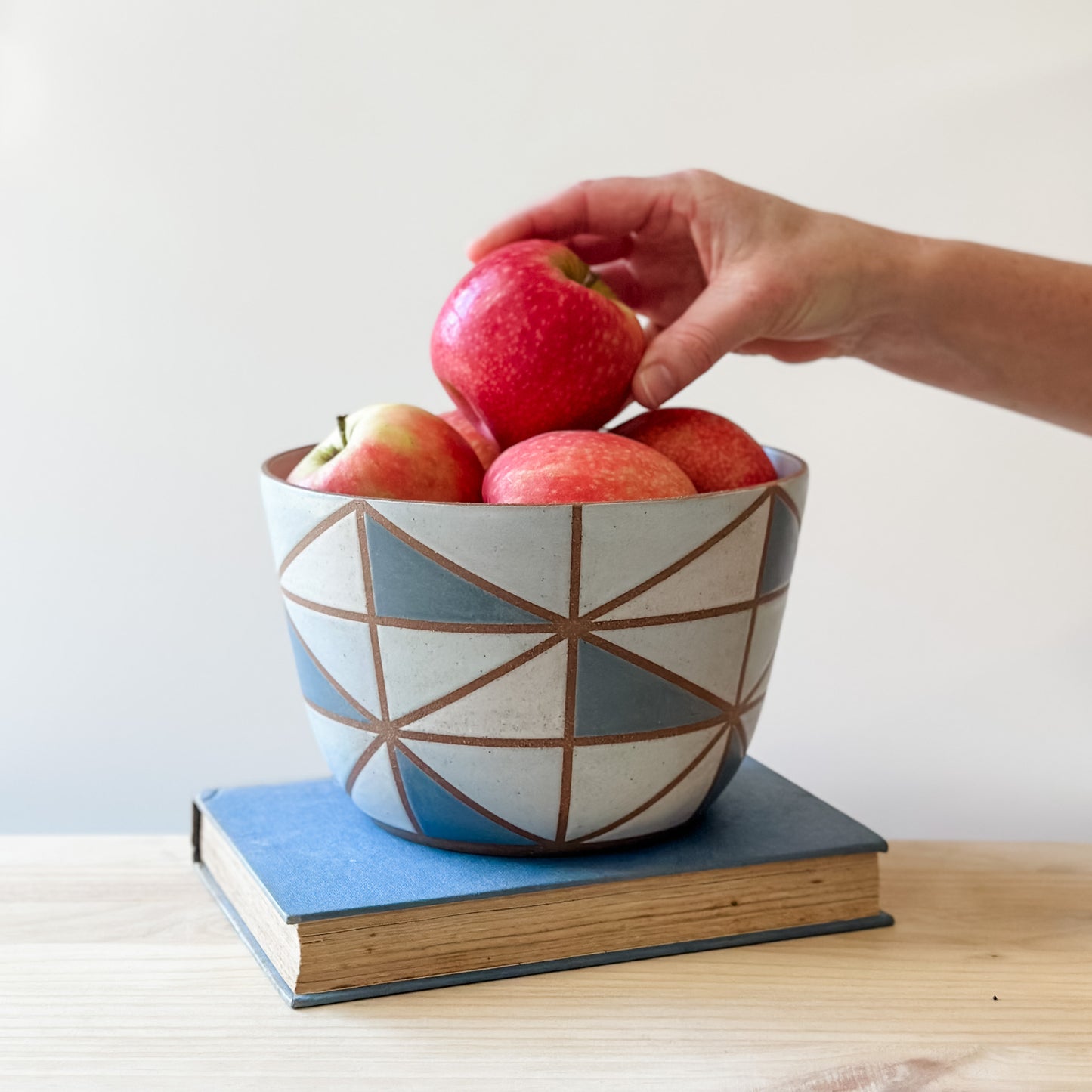 A geometric-styled handmade ceramic bowl sits on a book on top of a table. The ceramic bowl is hand-painted in dark blue, light blue, white and grey. A hand is placing fresh, red apples into the bowl.