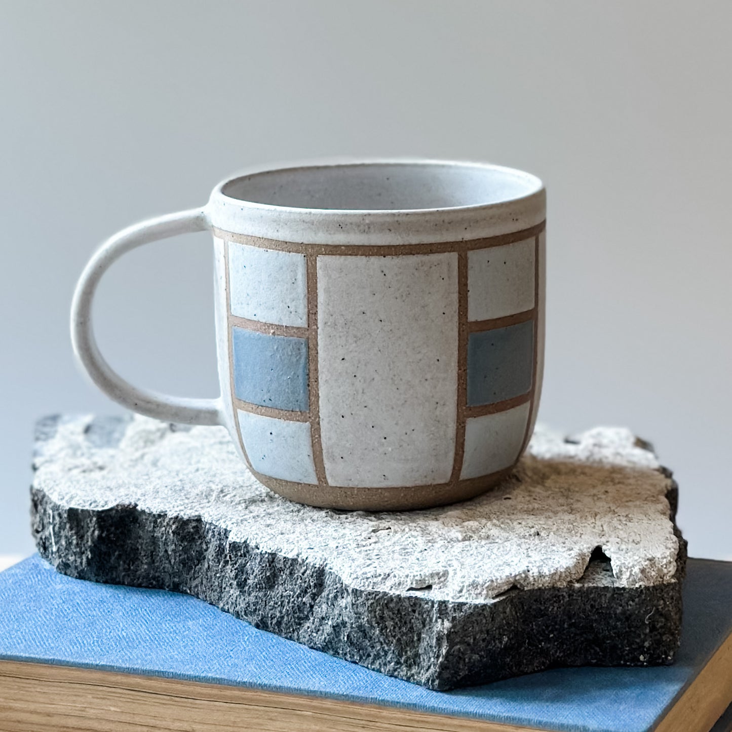A close-up view of a handmade ceramic mug with a geometric hand-painted design. The handmade mug sits on a tile and on a book. The mug is painted white, blue and grey.