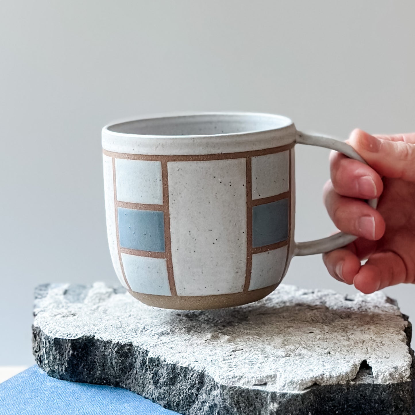 A handmade ceramic mug with a geometric hand-painted design. The handmade mug is being held above a tile. The mug is painted white, blue and grey.