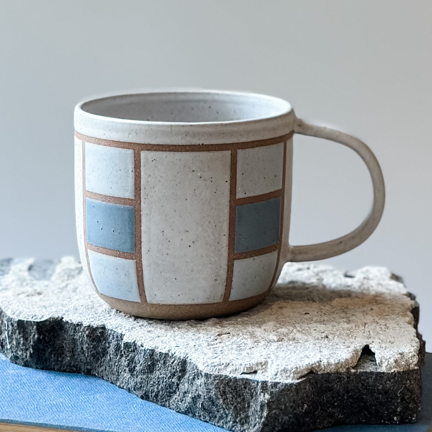 A close-up view of a handmade ceramic mug with a geometric hand-painted design. The handmade mug sits on a tile and on a book. The mug is painted white, blue and grey.