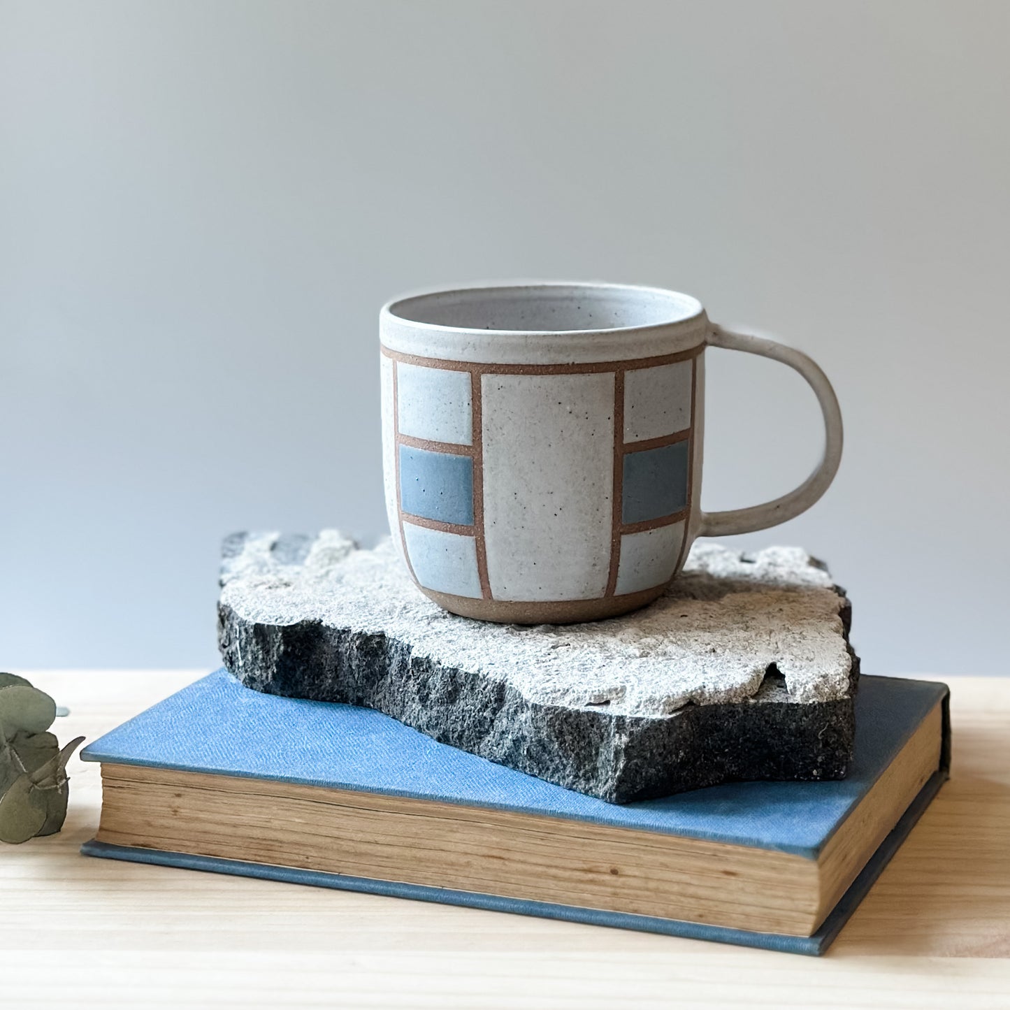 A handmade ceramic mug with a geometric hand-painted design. The handmade mug sits on a tile and on a wooden table. The mug is painted white, blue and grey.