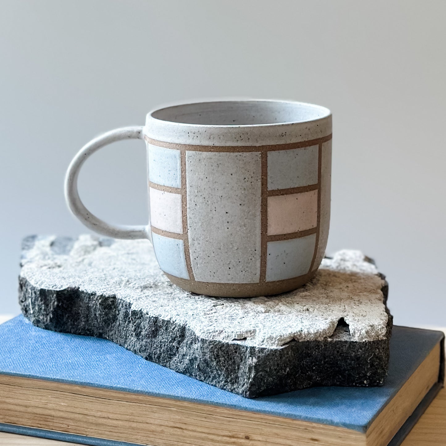 A close-up view of a handmade ceramic mug in a geometric design. The mug is painted white, pink and grey. The ceramic mug is sitting on a tile, a book and a wooden table.