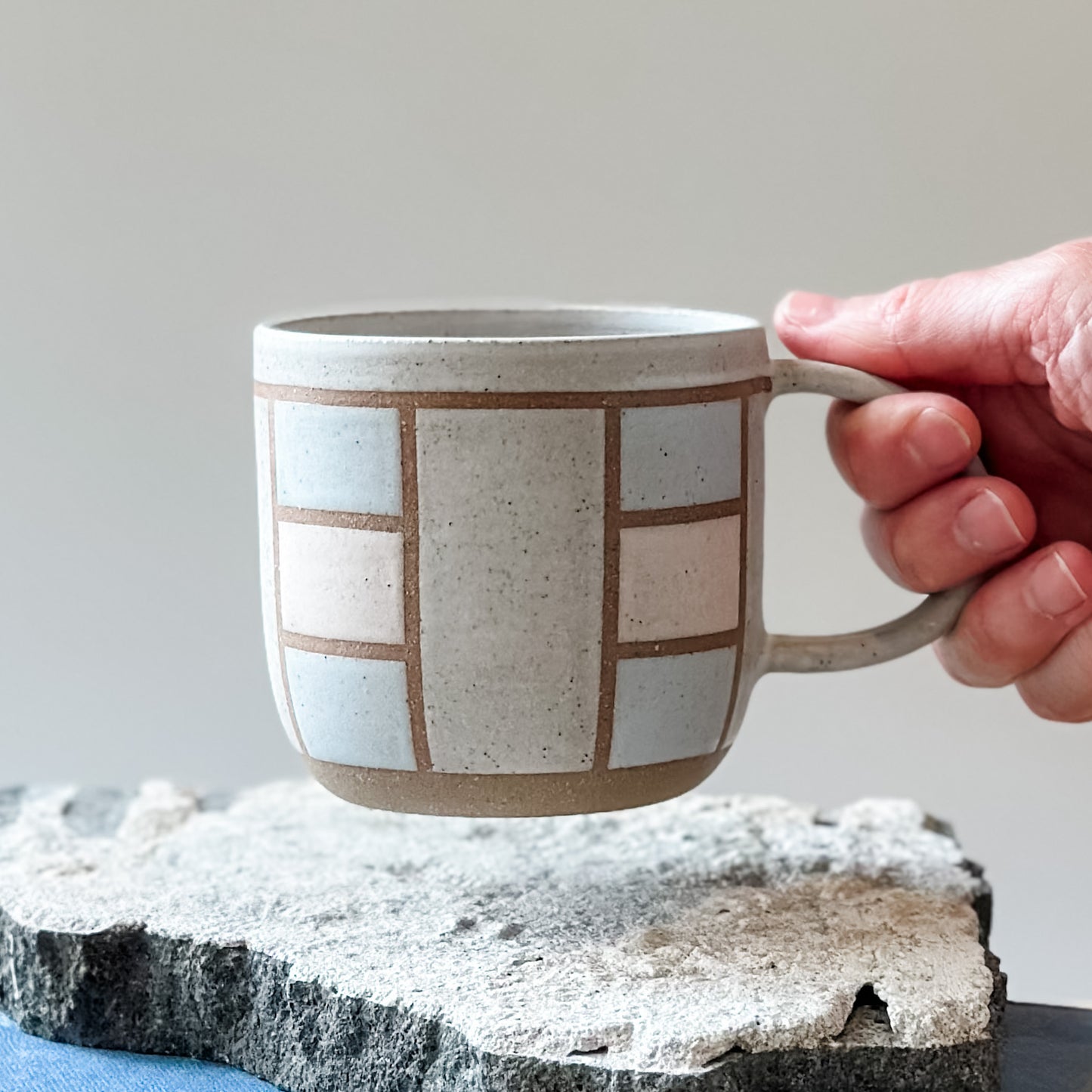 A handmade ceramic mug in a geometric design. The mug is painted white, pink and grey. The ceramic mug is being held above a tile by a hand.