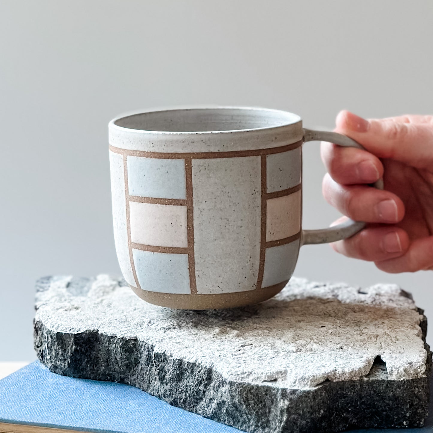 A handmade ceramic mug in a geometric design. The mug is painted white, pink and grey. The ceramic mug is being placed on a tile by a hand.