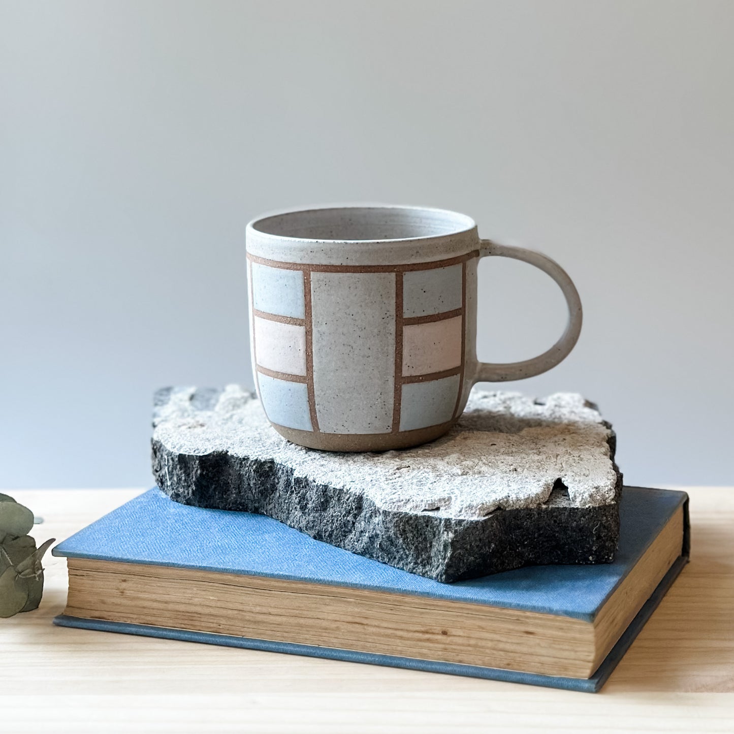 A handmade ceramic mug in a geometric design. The mug is painted white, pink and grey. The ceramic mug is sitting on a tile, a book and a wooden table.