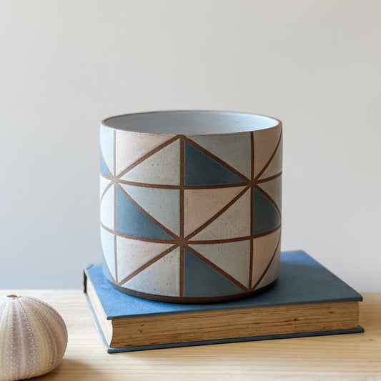 A geometric handmade ceramic planter pot sits on a book and wooden table. The planter pot is painted with dark blue, pink, grey and white glaze.