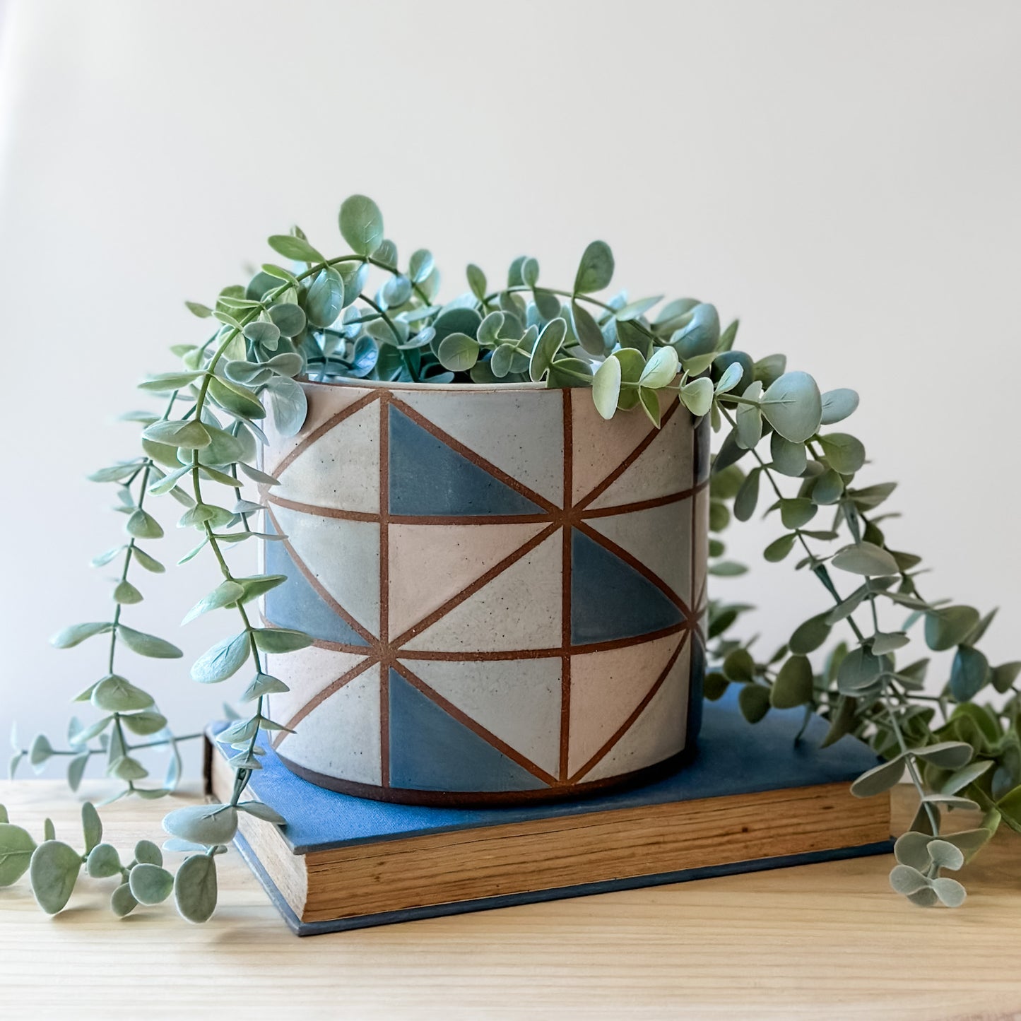 A geometric handmade ceramic planter pot sits on a book and wooden table. The planter pot is painted with dark blue, pink, grey and white glaze. The handmade planter has a hanging plant.