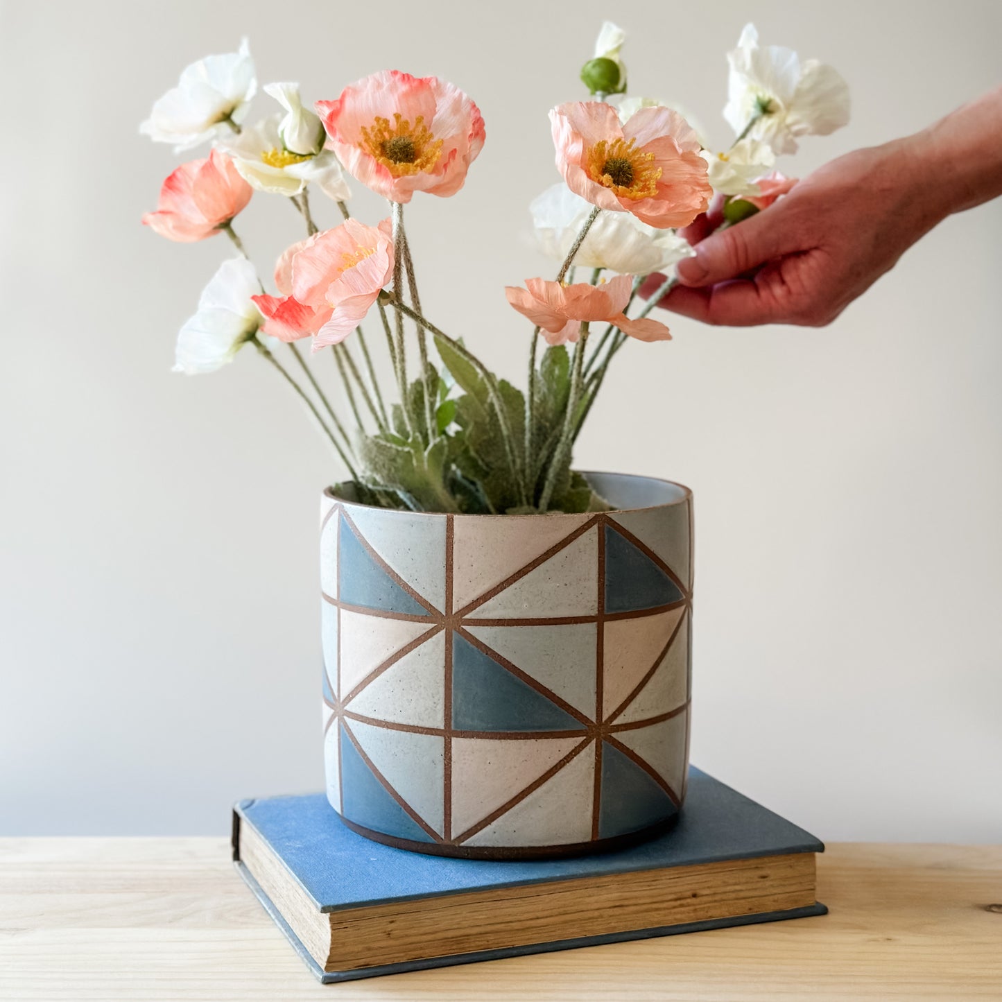 A geometric handmade ceramic planter pot sits on a book and wooden table. The planter pot is painted with dark blue, pink, grey and white glaze. A hand is arranging flowers in the ceramic planter.
